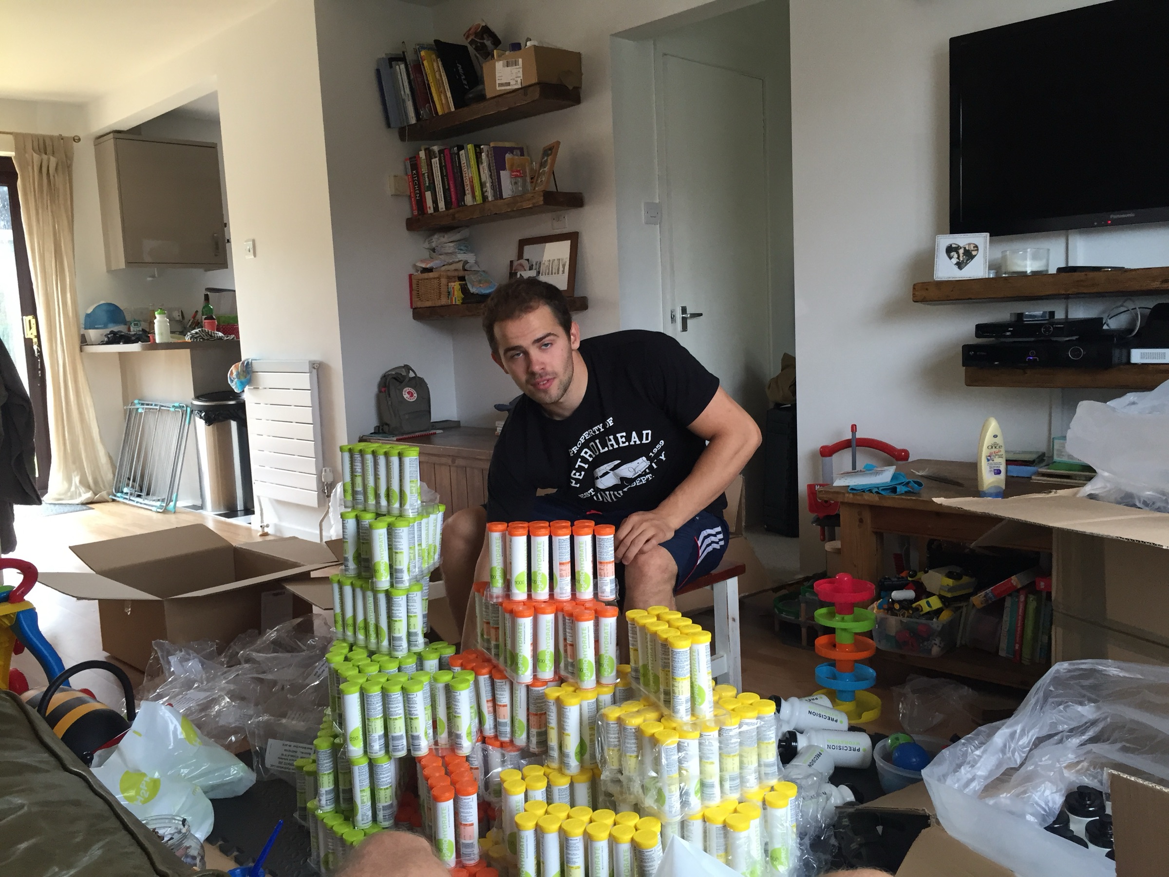 jonny tye labelling tubes of precision electrolytes by hand in andy blow's living room, surrounded by shipping boxes